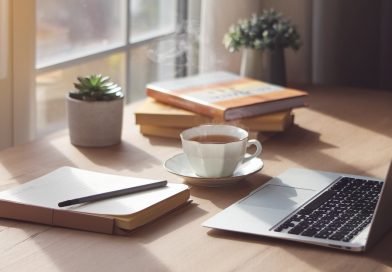 A peaceful and organised workspace featuring a cup of tea, a laptop, a journal, and a pen on a wooden desk with soft natural light and small potted plants in the background.