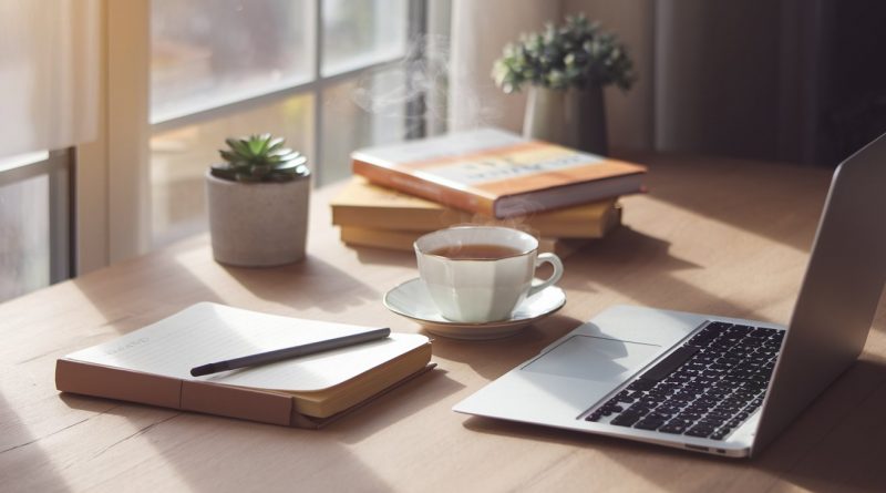 A peaceful and organised workspace featuring a cup of tea, a laptop, a journal, and a pen on a wooden desk with soft natural light and small potted plants in the background.