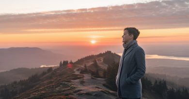 A person standing on a mountaintop at sunrise, gazing into the distance, with a winding path and a serene landscape of trees and hills below, symbolising self-discovery and introspection.
