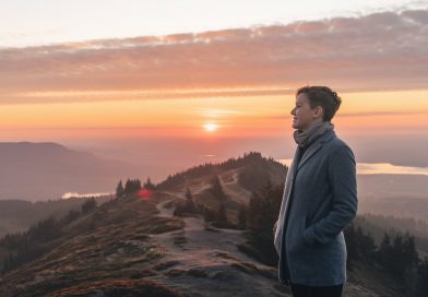 A person standing on a mountaintop at sunrise, gazing into the distance, with a winding path and a serene landscape of trees and hills below, symbolising self-discovery and introspection.