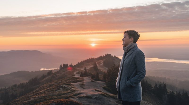 A person standing on a mountaintop at sunrise, gazing into the distance, with a winding path and a serene landscape of trees and hills below, symbolising self-discovery and introspection.