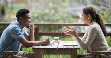 A serene outdoor setting featuring two individuals sitting at a wooden table, actively engaged in a warm and meaningful conversation, surrounded by lush greenery.