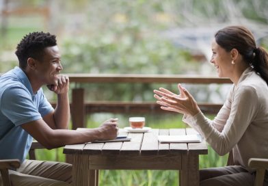 A serene outdoor setting featuring two individuals sitting at a wooden table, actively engaged in a warm and meaningful conversation, surrounded by lush greenery.