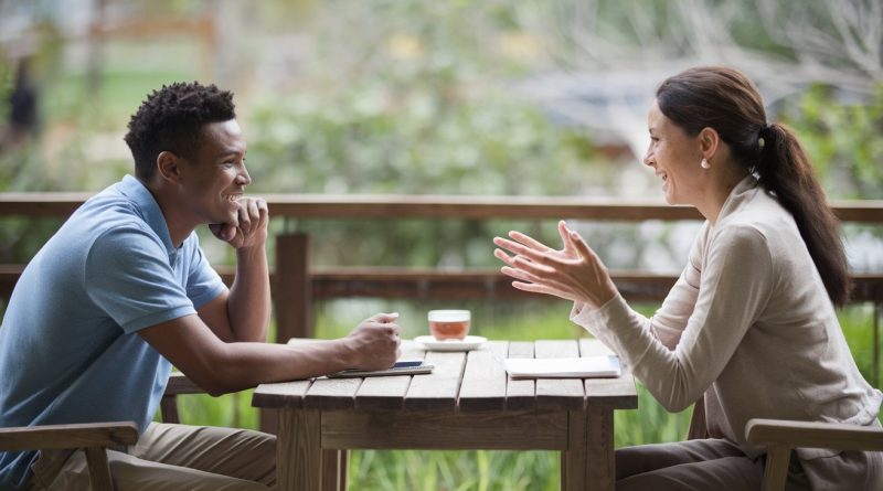 A serene outdoor setting featuring two individuals sitting at a wooden table, actively engaged in a warm and meaningful conversation, surrounded by lush greenery.